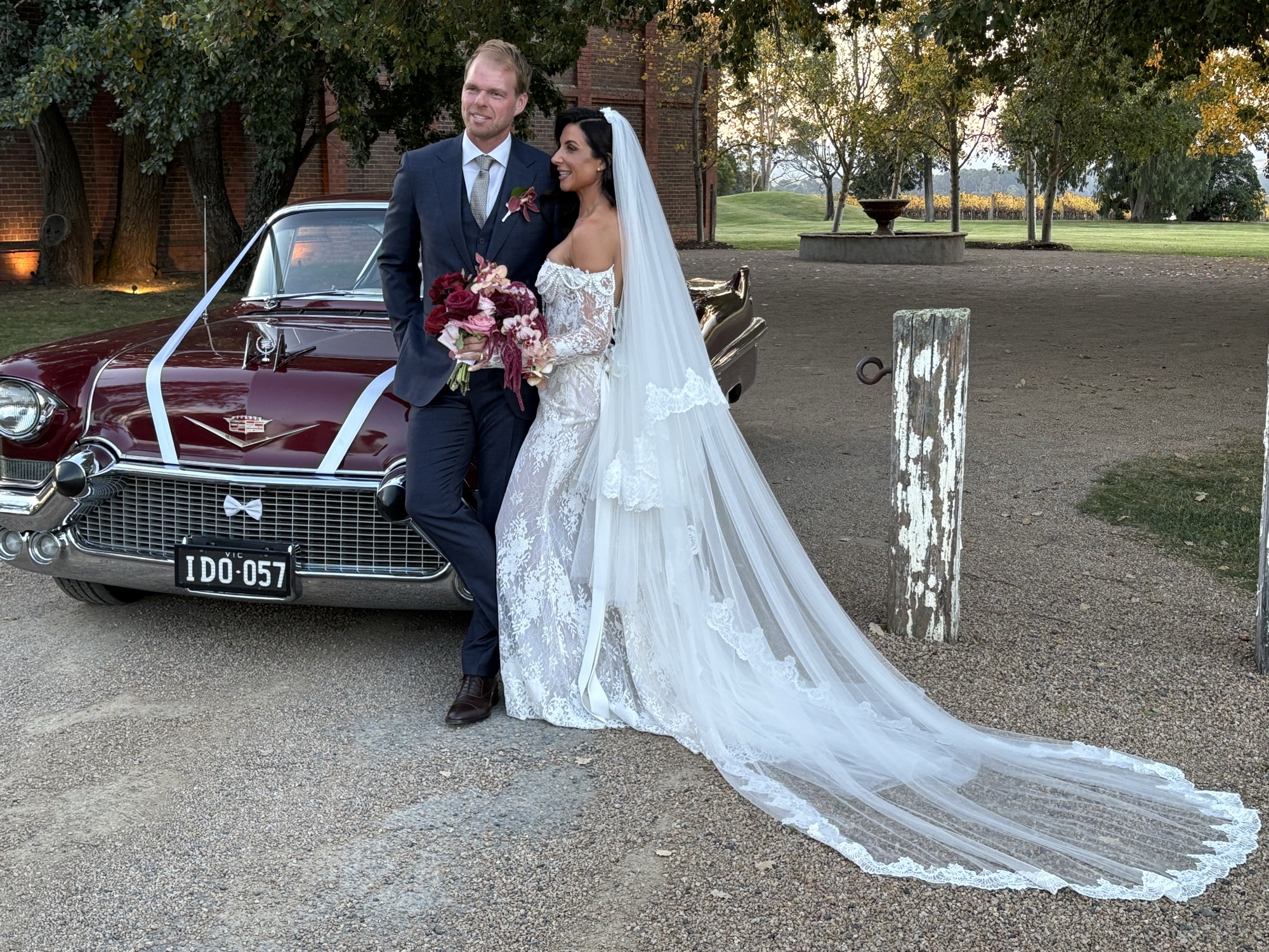 Bride and groom posing beside ELLIE Mae, the maroon classic Cadillac coupe, with the bridal veil flowing