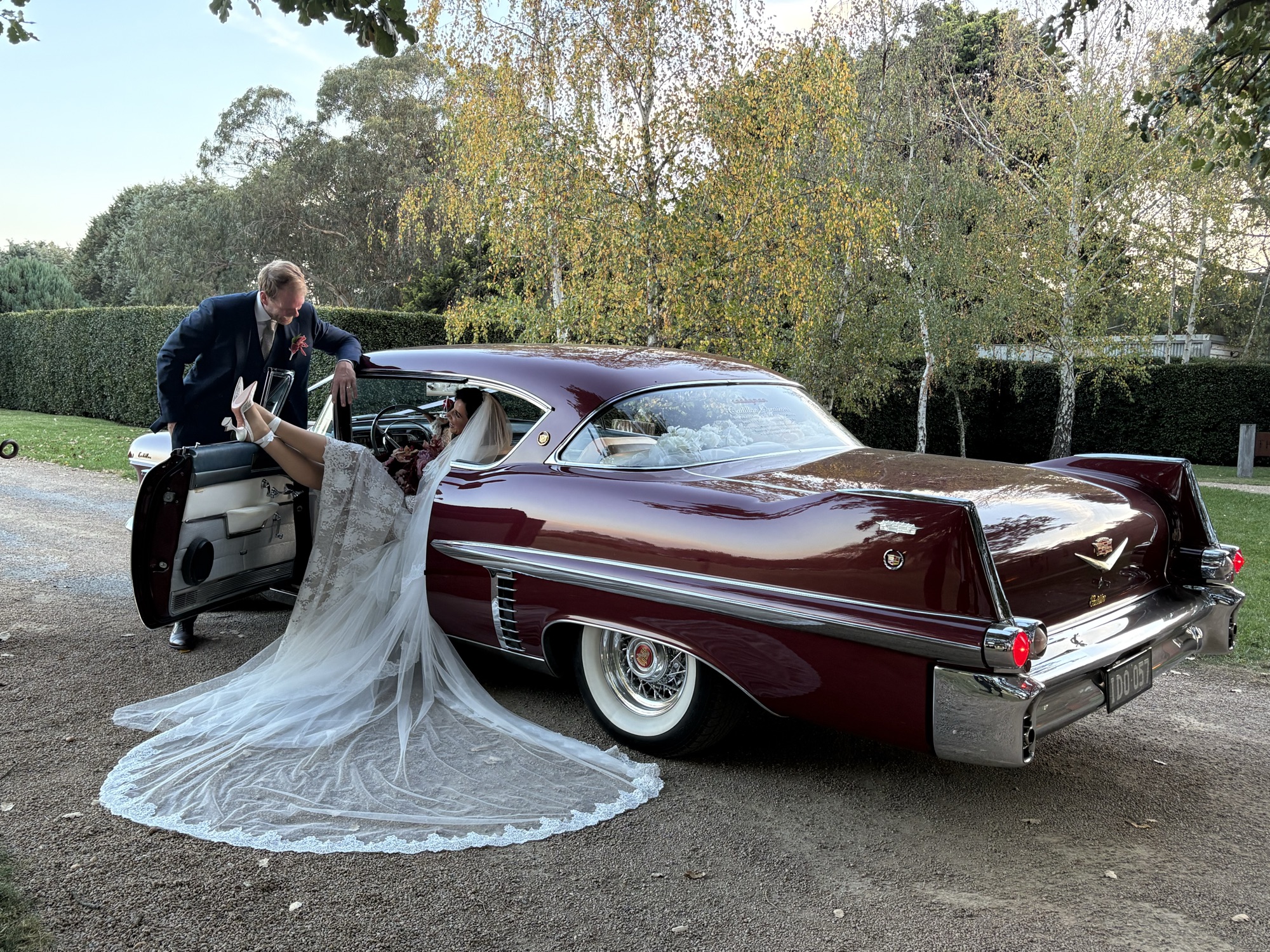 Bride seated in ELLIE Mae the maroon classic Cadillac coupe with her lace veil trailing out the open door