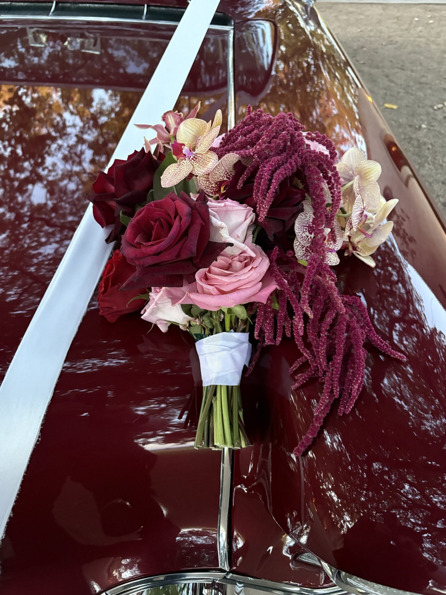 Bridal bouquet of roses and orchids resting on the maroon bonnet of ELLIE Mae, Caddyman's classic Cadillac