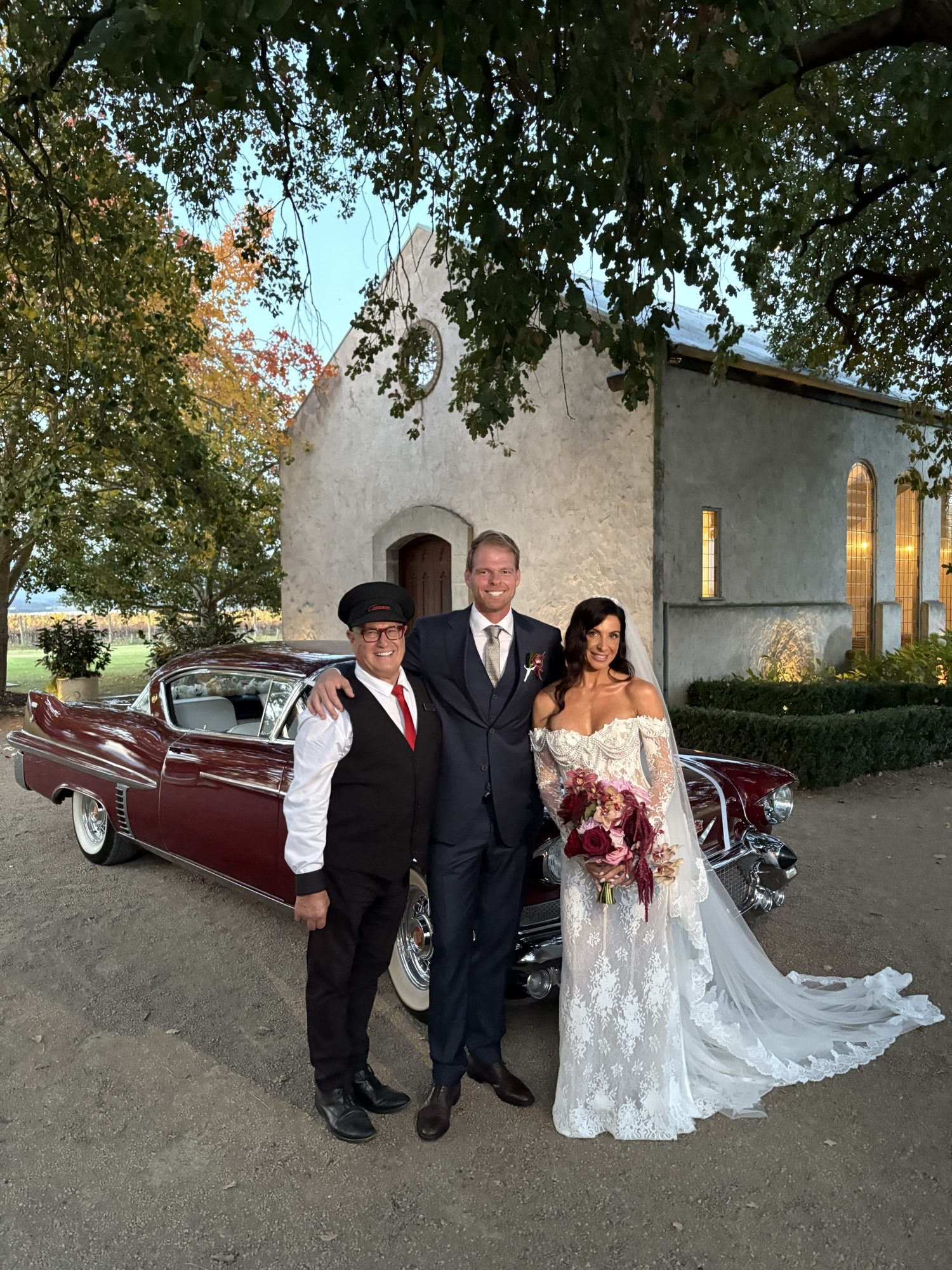 Bride and groom with their Caddyman chauffeur in front of ELLIE Mae, the maroon classic Cadillac coupe