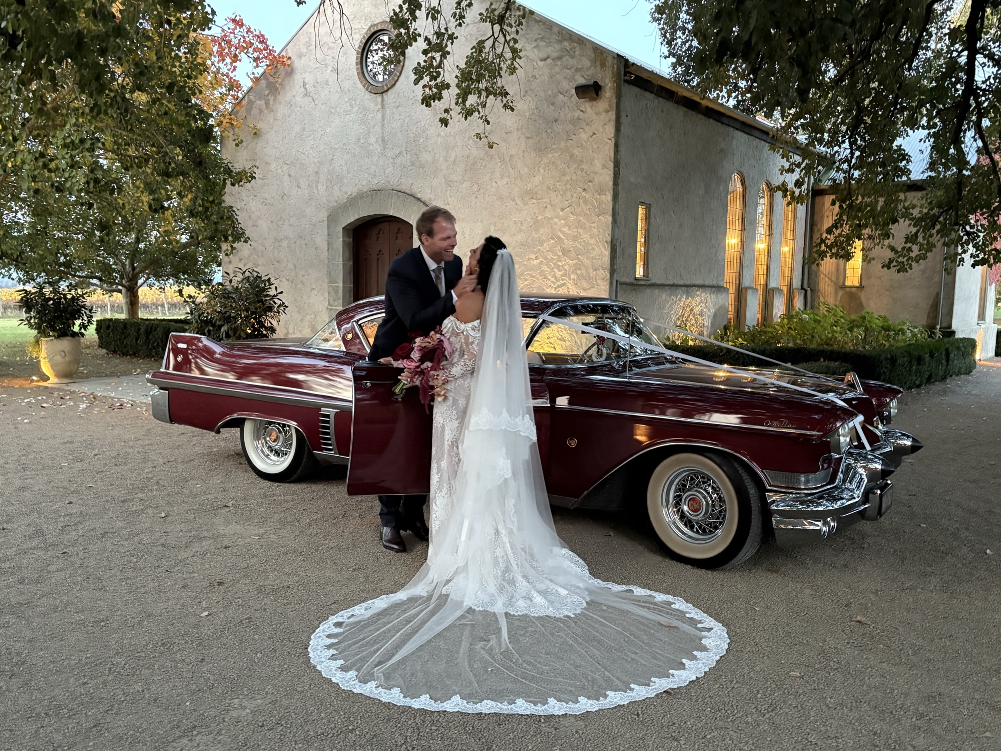Bride and groom kissing beside ELLIE Mae, the maroon classic Cadillac coupe, outside the chapel