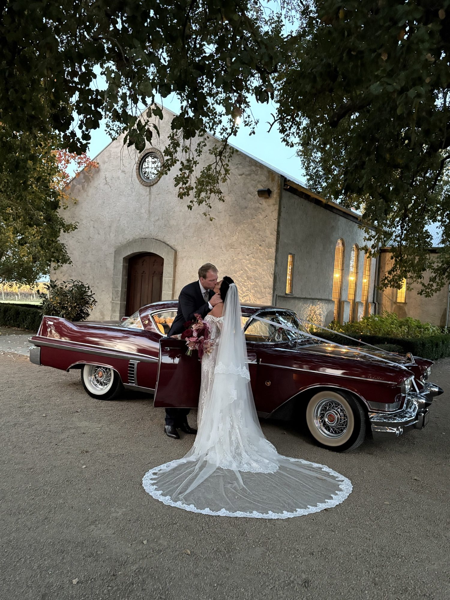 Newlyweds embracing beside ELLIE Mae the maroon classic Cadillac coupe, the bridal veil spread across the gravel