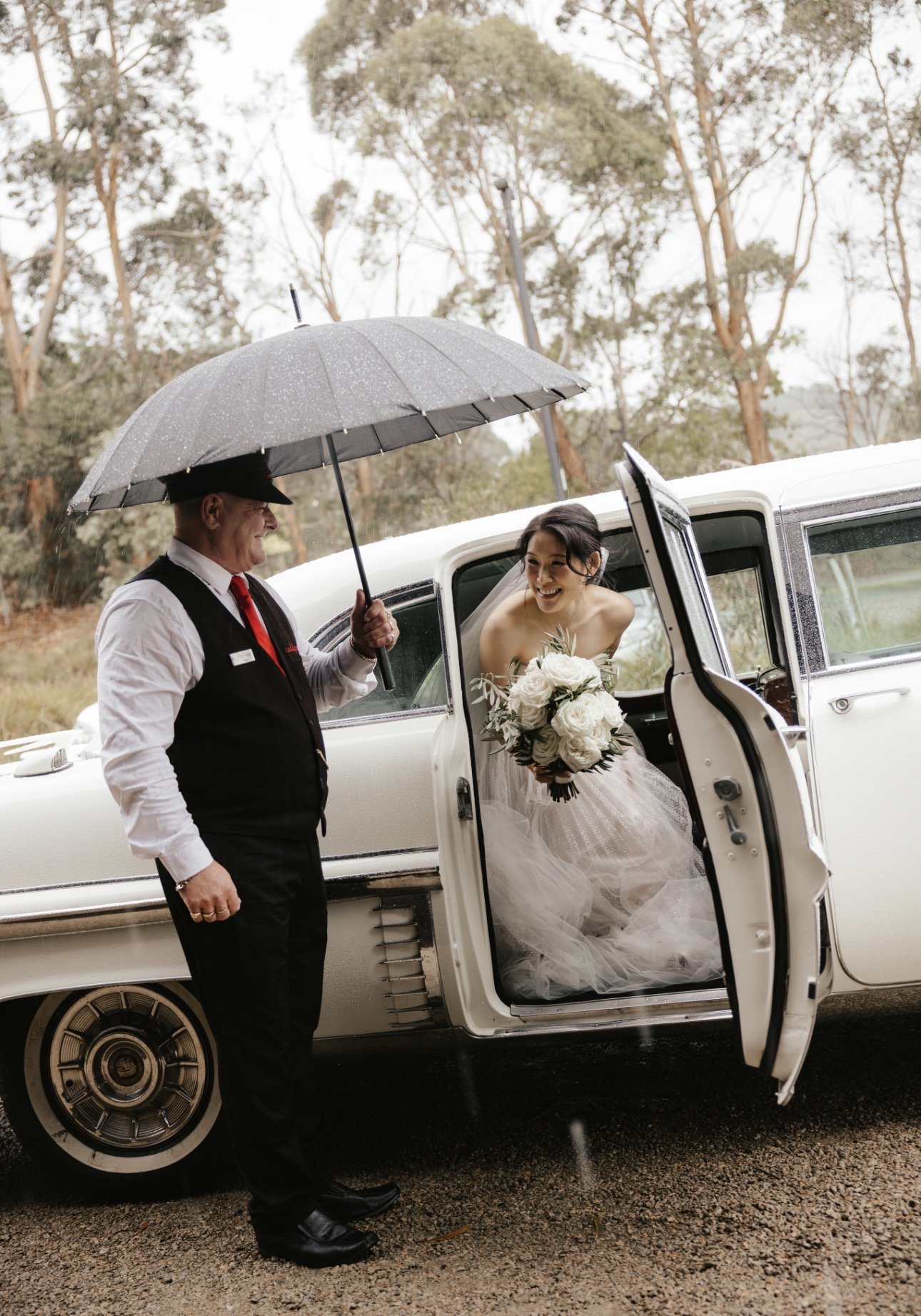 Chauffeur holding an umbrella for a bride stepping out of The Duchess, Caddyman's white 1957 Cadillac Series 75 wedding limousine, at a Melbourne wedding