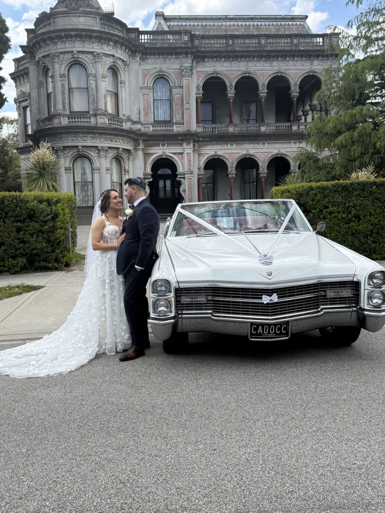 Bride and groom beside the front of Queen B, Caddyman's white 1966 Cadillac Coupe De Ville convertible, outside a historic Melbourne mansion