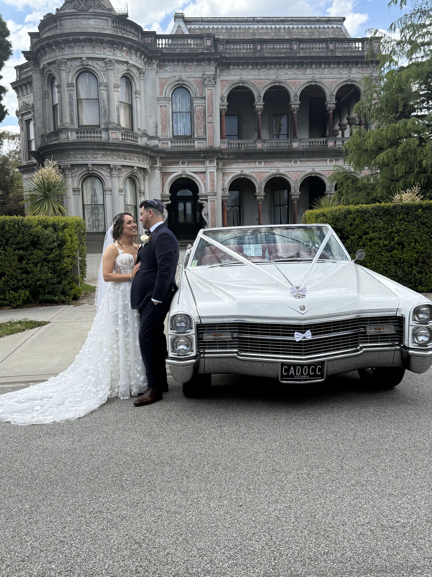 Bride and groom beside the front of Queen B, Caddyman's white 1966 Cadillac Coupe De Ville convertible, outside a historic Melbourne mansion