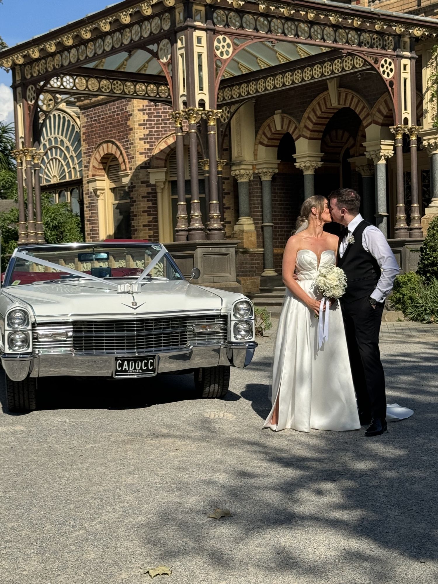 Bride and groom kissing beside Queen B, Caddyman's white 1966 Cadillac Coupe De Ville convertible, outside an ornate Melbourne mansion