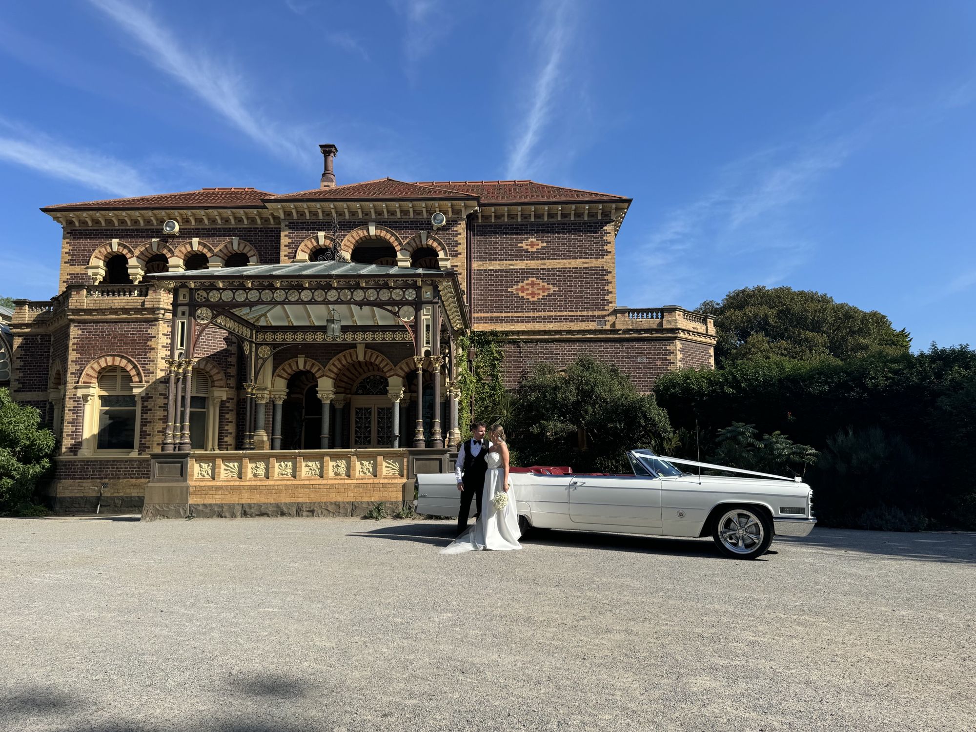 Queen B, Caddyman's white 1966 Cadillac Coupe De Ville convertible, parked in front of a historic mansion with the bride and groom
