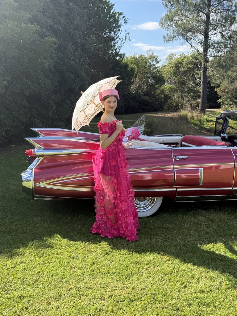 Guest in a pink dress with a parasol beside Rosie, Caddyman's 1959 Rose Pink Pearl Cadillac convertible, on the lawn