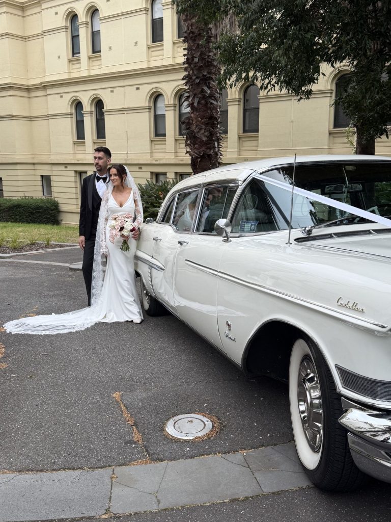 Bride and groom beside The Duchess, Caddyman's white 1957 Cadillac Series 75 wedding limousine, outside a heritage city building