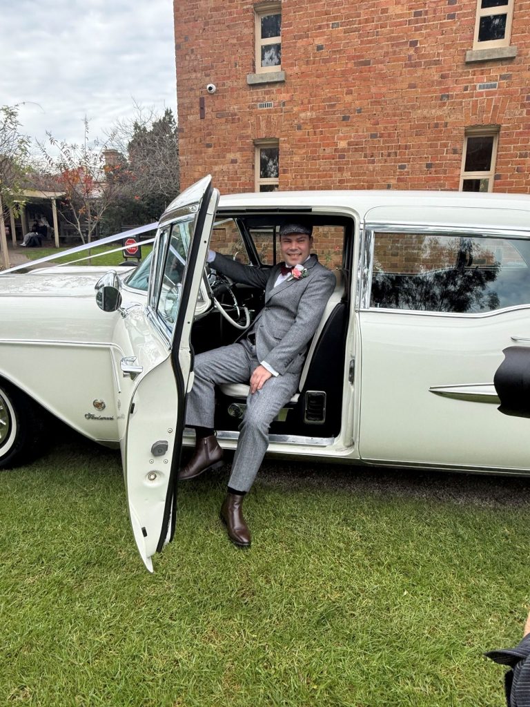 Groom seated in the rear doorway of The Duchess, Caddyman's white 1957 Cadillac Series 75 wedding limousine with ribbons on the bonnet